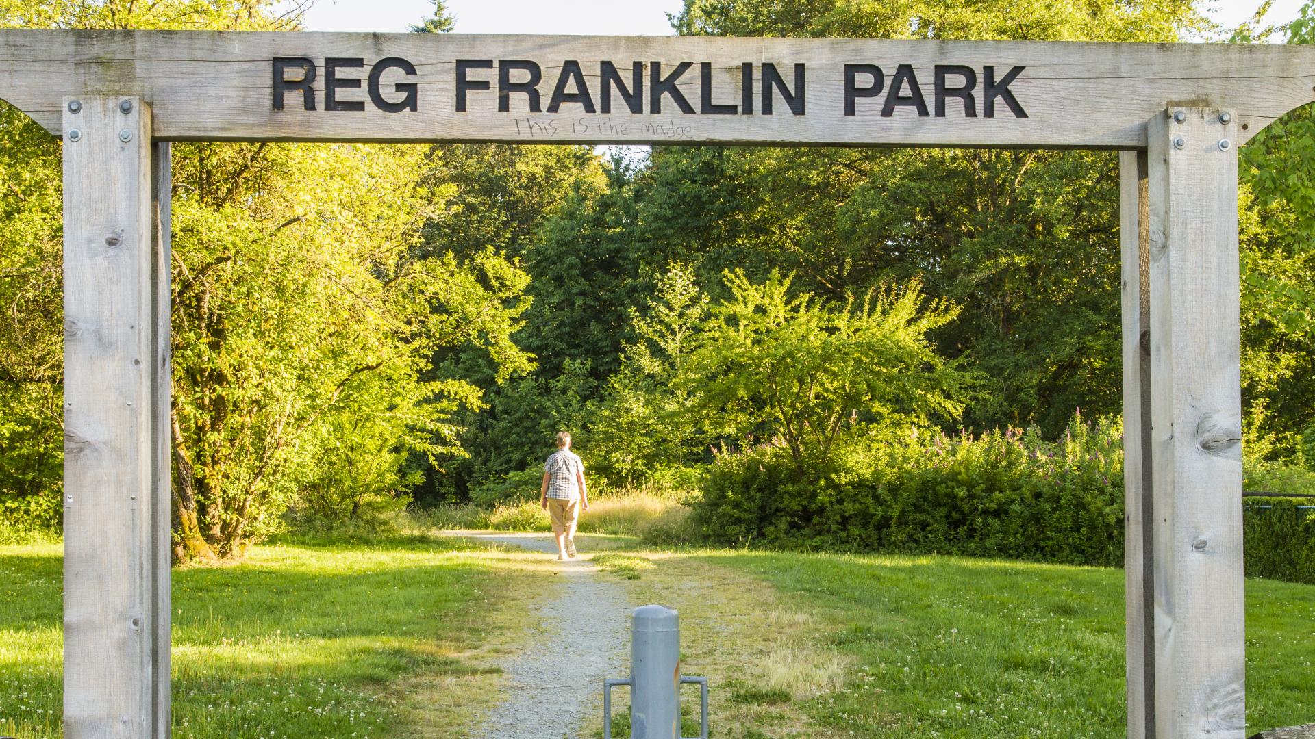 A wood archway with "Reg Franklin Park" across the top beam. The archway is over a gavel pathway the leads into a forested area.